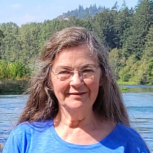 Smiling woman with long gray hair and glasses, wearing a blue shirt, standing outdoors by a lake with trees in the background.